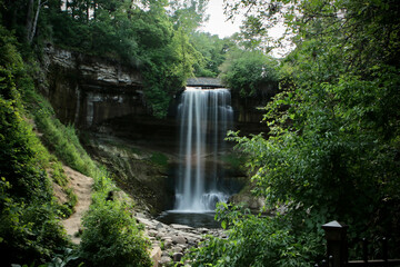 A graceful waterfall cascades from a rocky cliff into a serene pool, framed by dense green foliage at Minnehaha Falls in Minnesota.
