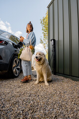 Young woman plugs a charger into electric vehicle standing with her cute white dog near her house...