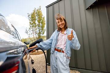 Portrait of a young woman plugging a charger into electric vehicle near her house outdoors. Concept...