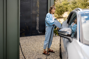 Young woman plugging a charger into electric vehicle, carrying bag with fresh vegetables near her...