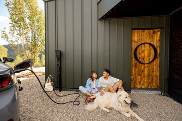 Lovely couple sitting with their dog near a modern house, electric car charges nearby. Concept of...
