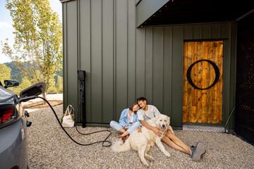 Lovely couple sitting with their dog near a modern house, electric car charges nearby. Concept of love, modern living and wellbeing