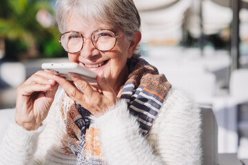 Portrait of smiling senior gray haired woman sitting outdoors in a sunny day recording a message on mobile phone. Old people and modern technology. Serene retirement lifestyle concept