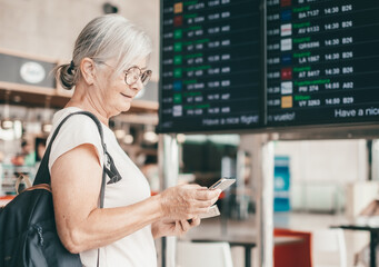 Smiling senior woman in airport looking at timetable schedule to check flight departure gate....