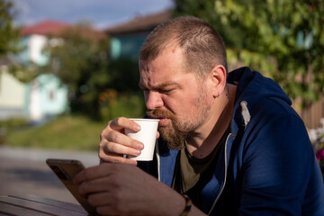 A bearded man sips coffee from a white cup while looking at his smartphone, seated outdoors on a...