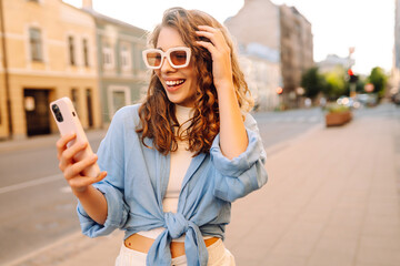 Picture of pretty young woman staying on the street holding phone in hands. Blogging, technology,...