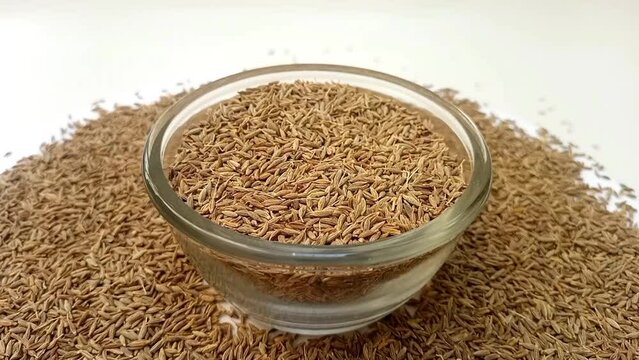 Cumin seeds in a glass bowl spices spilled around it, rotating on white background, center frame, zooming in, close up, herbs and spices.