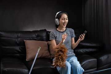 Smiling woman dusting with feather duster and listening to music on headphones. Concept of...