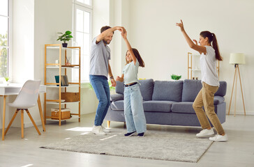 Joyful people having fun at home. Happy vibrant family dancing to lively music together. Young energetic mother, father and child having party and dancing in modern spacious cozy living room interior