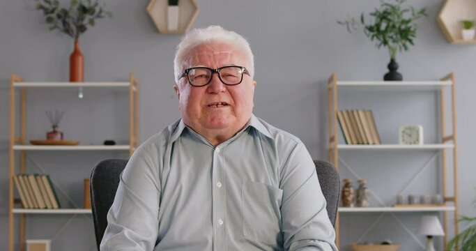 Elderly man shares his life stories on camera with expressive gestures and emotions. Caucasian pensioner in shirt and glasses in room sits in front of camera and cheerfully tells stories. 4k footage.