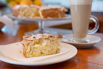 Tasty slice of cake on a plate accompanied by a tall glass of a cafe latte, delicious dessert featuring an apple and almond sponge cake topped with sugar and nuts flakes and served with a large coffee