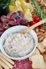 Close-up of a creamy dip in a white ceramic bowl, surrounded by a colorful assortment of appetizers on a charcuterie board such as red grapes, cherry tomatoes, row almonds, mini toast slices and more