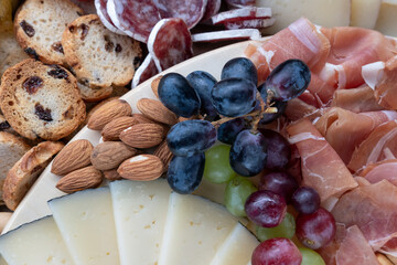 Appetizing cold spread with several cured meats and sliced cheeses, accompanied by tasty bread products, fresh fruit and dried nuts, close-up of a charcuterie board prepared for a gathering or a party