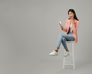 full lenght shoot of happy excited asian woman sitting on stool holding mobile phone and giving...