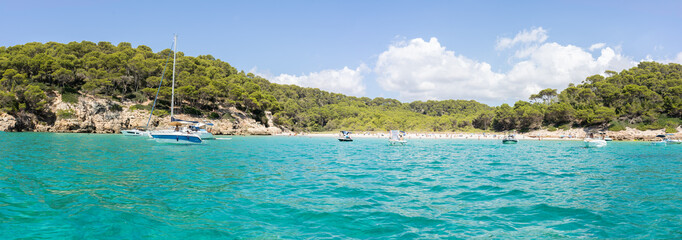 View of Cala Turqueta in the south of the island of Menorca as seen from offshore, with people on the beach sunbathing and swimming and boats at anchor