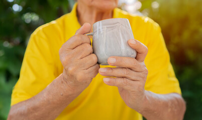 Hands of old man wearing yellow t-shirt holding cup of coffee over nature background.