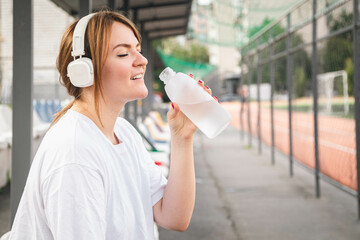 Woman in white headphones drinks water after training at the stadium, concept of outdoor sports, water balance, copy space.