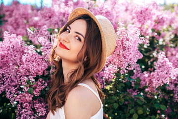 Young beautiful smiling woman in trendy summer clothes. Sexy carefree model posing in the street. Positive model near blooming flower lilac bush beaming. Summertime, pink colours. Red lips, straw hat