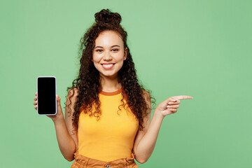Young happy woman wear yellow tank shirt top hold in hand use mobile cell phone with blank screen workspace area point index finger aside isolated on plain pastel green background. Lifestyle concept.
