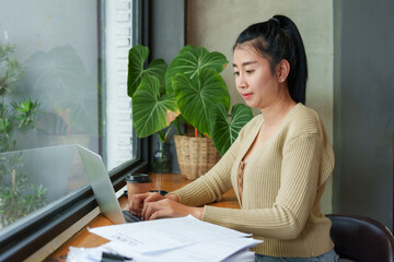 Asian female in beige sweater focused on laptop, paperwork beside, coffee cup, natural light, green...