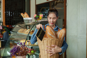 Asian woman female in blue shirt, apron, presenting fresh bread in a basket, cheerful in a rustic bakery setting. happiness of opening a coffee shop in the morning small family business