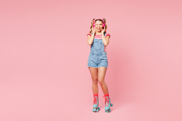 Full body young happy woman wear red t-shirt denim overalls casual clothes blue rollers rollerblading listen music in headphones wink isolated on plain pink background. Summer sport leisure concept.