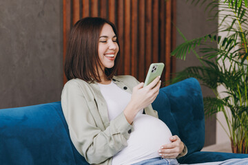 Young smiling pregnant woman with belly wear casual clothes hold use mobile cell phone sit on blue sofa couch stay home hotel flat rest relax spend free time in living room indoor. Maternity concept.