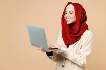 Young happy Arabian Asian Muslim IT woman wear red abaya hijab suit clothes hold use work on laptop pc computer isolated on plain beige background studio. UAE middle eastern Islam religious concept.