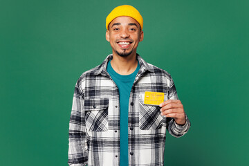 Young smiling fun man of African American ethnicity he wear shirt blue t-shirt yellow hat hold in hand mock up of credit bank card isolated on plain green background studio portrait Lifestyle concept