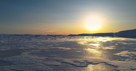Fly over sunset frozen ocean in Antarctica. Sun warm glow reflect on icy surface. Antarctica travel background and exploration. Discover the beauty of South Pole. Desert white land of snow drone shot.