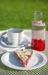 A piece of homemade cake with red and black currant and crumb on a white plate, a cup of coffee with saucer and a teaspoon, currant lemonade and a striped tea towel on a white table in the garden