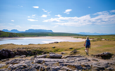 A female hiker and their dog walking towards the remote sandy beach and blue turquoise water of Achnahaird Bay with distant views of Sgorr Tuath and Ben More Coigach in the Scottish Highlands