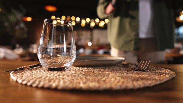 Faceless man in casual clothes and black gloves placing empty whiskey glass at table with plate and cutlery. Waiter complementing serving with container for drink before client arriving.
