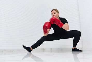 European woman fighter boxer in red boxing gloves, doing stretching exercises on legs,warming up,...