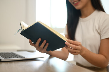 Peaceful young Asian woman in casual clothes reading book on desk at home
