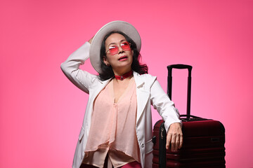 Confident middle age woman in sunglasses and straw hat standing with suitcase on pink background