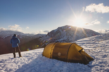 A solitary camper stands near a yellow tent, overlooking snow-covered mountains as the sun rises.