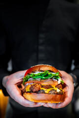 Close-up of hands presenting a gourmet burger with fresh greens, tomato, cheese, and a juicy patty...