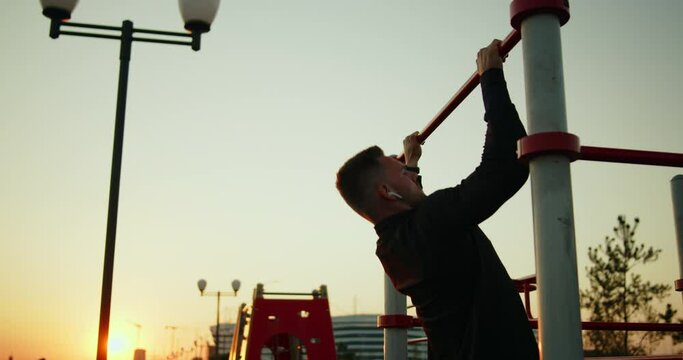 Young athletic man training outside doing pull-ups on a street sports ground at sunset. Outdoor workout exercises. Active healthy lifestyle, wellbeing and body care. Slow motion medium shot.