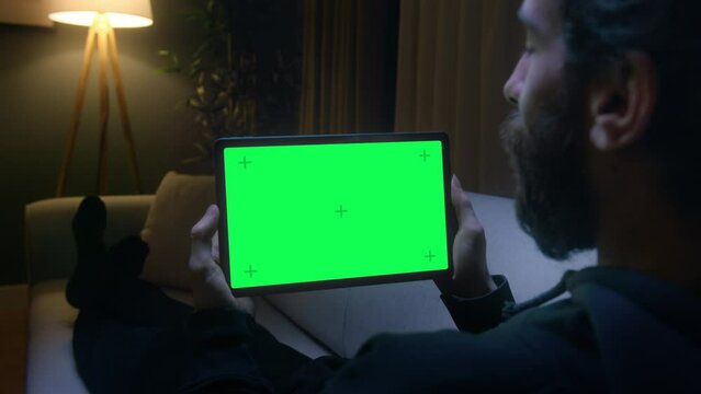View from shoulder of man lying on couch using tablet with chroma key green screen. Man watching series or video at home at night.