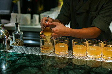 Barman pouring fresh juice into the glasses with whiskey and ice cubes for finishing cocktail on the bar counter