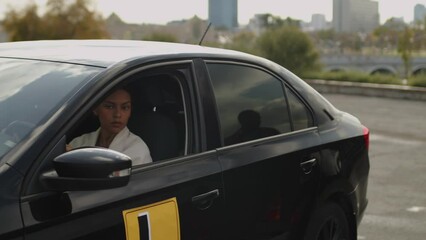 Pan of smiling young African American woman sitting in black learners car passing driving test under supervision of experienced male instructor holding clipboard