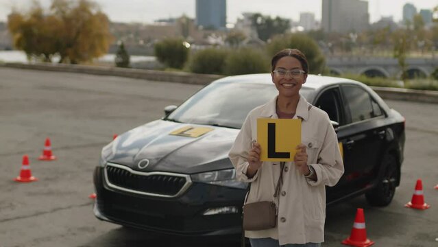Medium portrait of young smiling African American woman wearing glasses standing at parking area near learners auto holding L plate ready for driving lesson