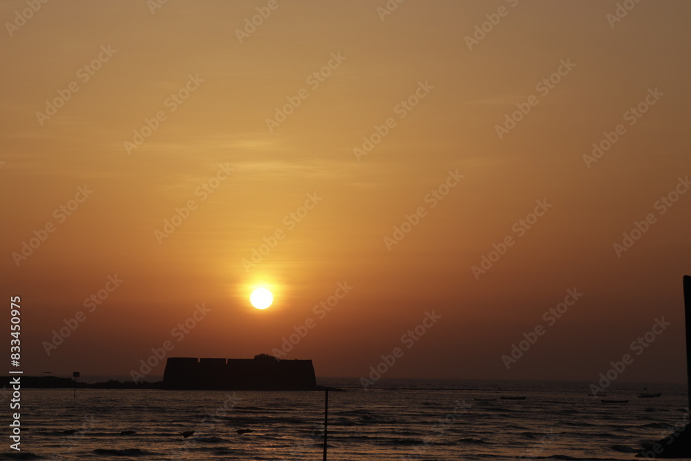 Poster Red sun setting on a beach with a ship in the water at dusk