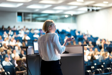 Female speaker giving a talk on corporate business conference. Unrecognizable people in audience at...