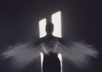 An angel with wings standing in the darkness in front of a door of light. 