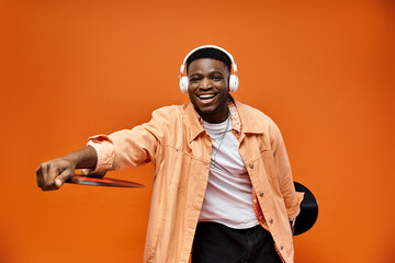 Stylish black man wearing headphones holding record on orange background.