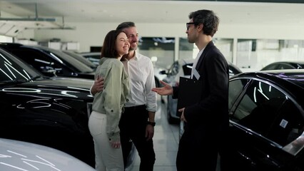 Happy couple a middle-aged man with gray hair together with his brunette wife is having fun talking with a car dealership assistant in a business suit about cars in a car showroom - Powered by Adobe