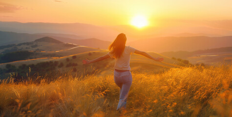 A woman is running on the grassy hill, with her arms outstretched and smiling as she looks at the sun setting behind her in warm tones.