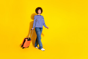 Full length photo of positive woman dressed striped shirt go empty space look at offer hold valise isolated on yellow color background